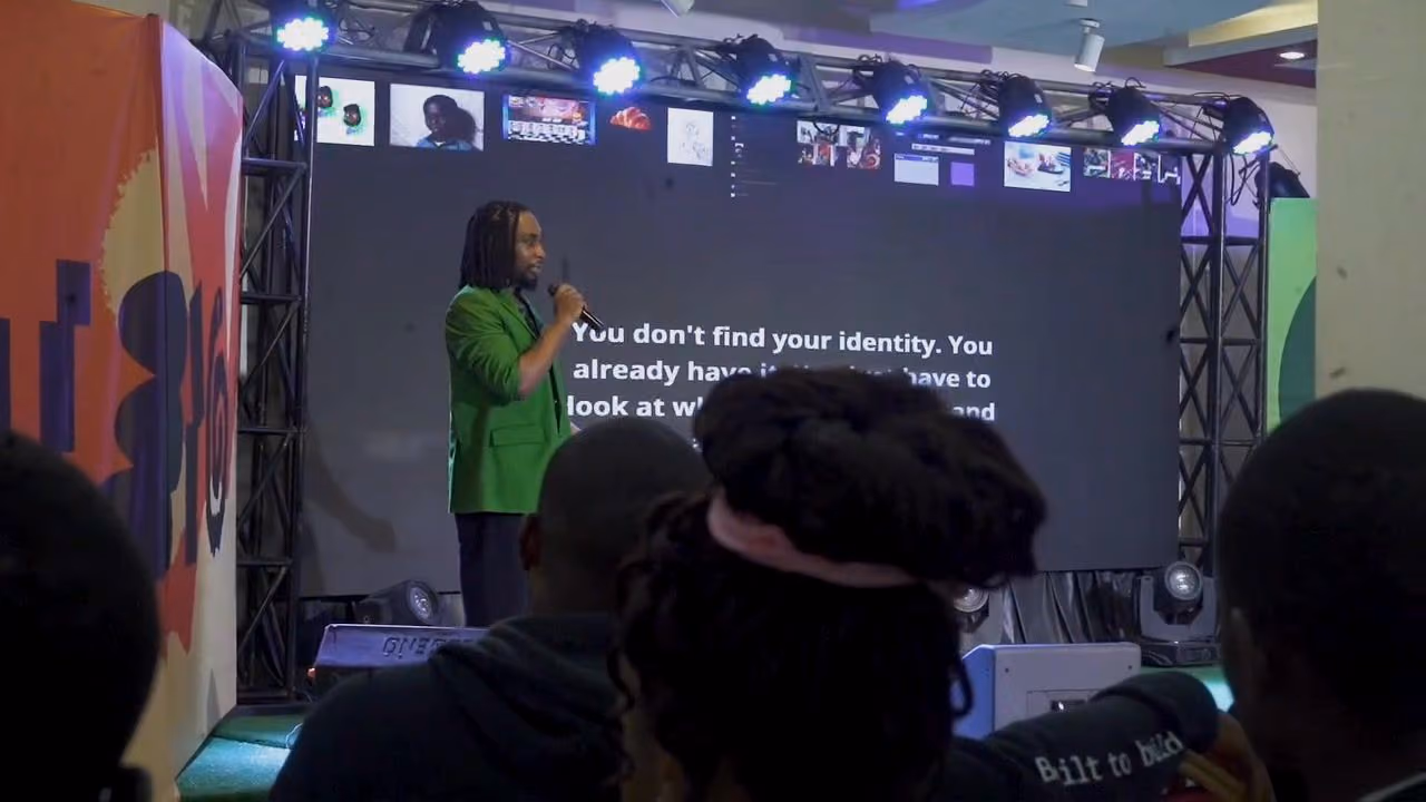 A speaker in a green blazer talks on stage beside a large quote slide while the audience watches