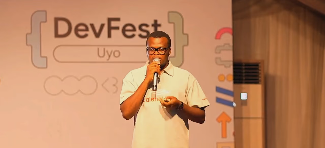 A speaker in a cream polo talks into a microphone on the DevFest Uyo stage