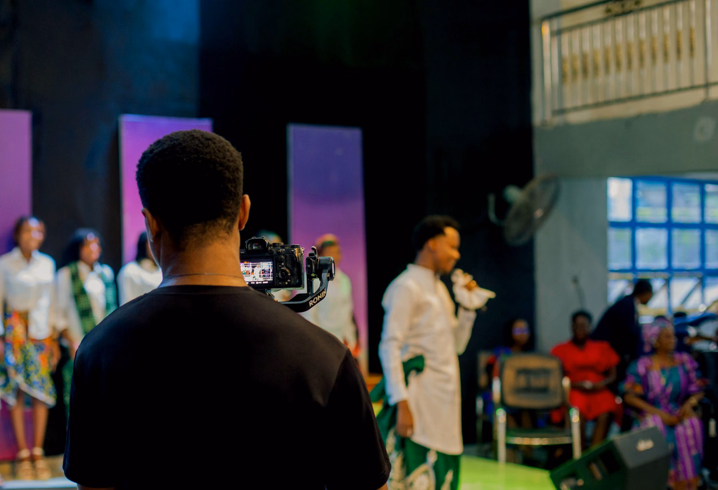 Camera operator seen from behind filming a stage lit in purple while the congregation stands